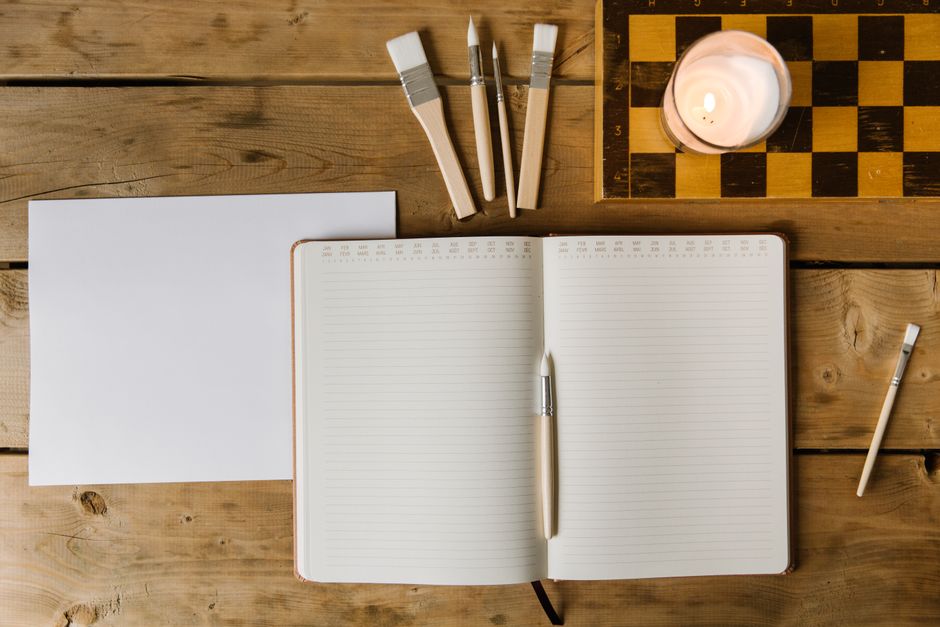 Top view of an open notebook with paintbrushes and lit candle on a rustic wooden desk.